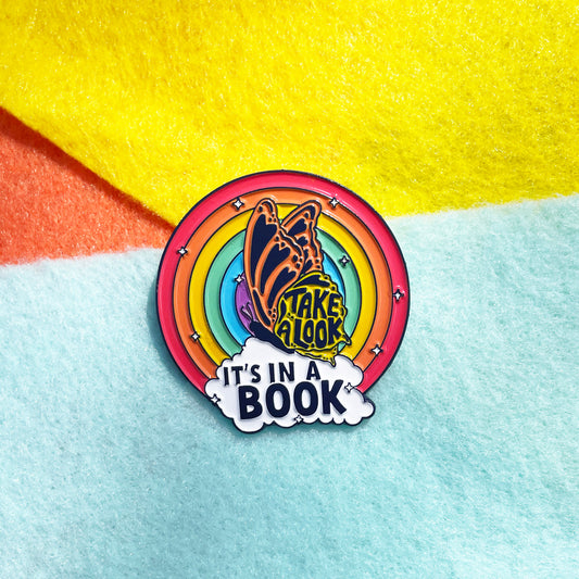 Reading Rainbow inspired colorful enamel pin with a rainbow and book design, featuring the text take a look 'IT'S IN A BOOK' with a butterfly